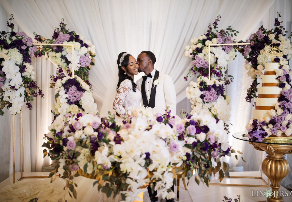 African wedding couple beside a purple and white floral arch with a tiered cake